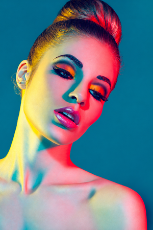 Woman with bold orange and yellow eye makeup and hair bun illuminated by colorful red and yellow studio lights.
