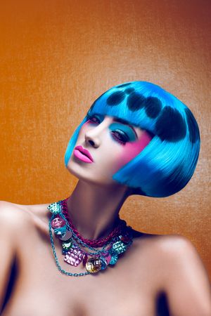 Model with bold blue bob wig featuring dark spots, pink makeup, and colorful statement necklace on orange background.