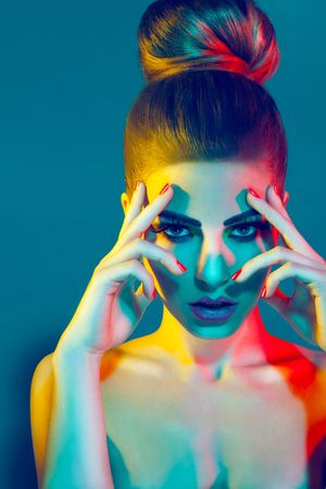Model with hair bun poses with hands near face under dramatic colorful studio lighting on teal background.