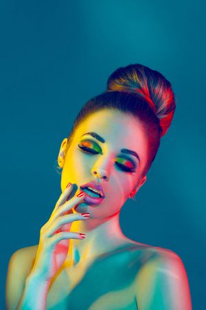 Fashion model with hair bun and colorful eye makeup poses sensually under vibrant colored studio lights.