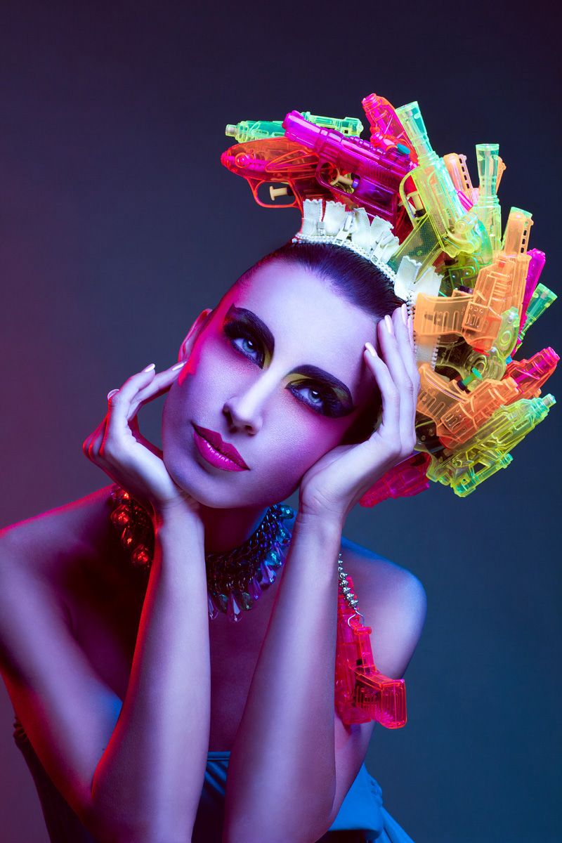 Creative fashion model wearing elaborate headpiece made of colorful toy water guns under pink studio lighting.