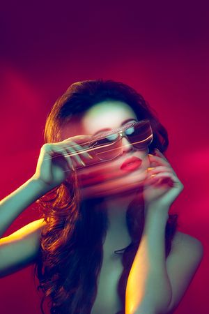 Woman with long wavy hair adjusting sunglasses with motion blur effect under dramatic red and yellow lighting.