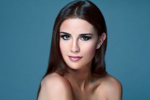 Beautiful brunette woman with sleek straight hair and subtle makeup poses against soft blue background.