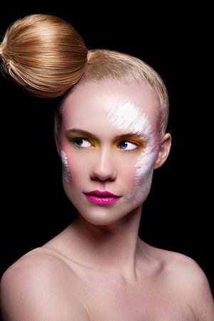 Woman with artistic white paint strokes on face, colorful eye makeup, pink lips, and sleek blonde hair bun on black background.