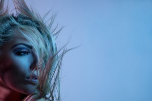 Blonde woman with blue-tinted hair blowing dramatically across her face under moody blue and red studio lighting.