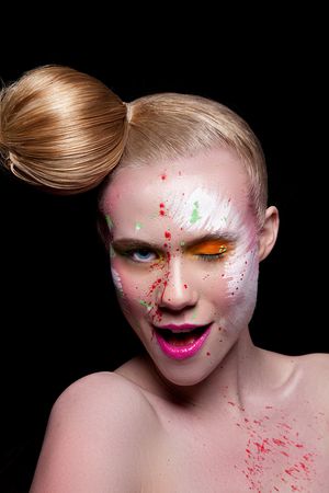 Woman with bold artistic makeup, colorful paint splatter on face, hair bun, and pink lips on black background.
