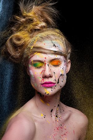 Woman lying down with vibrant multicolored paint splashes across her face and hair in artistic beauty portrait.
