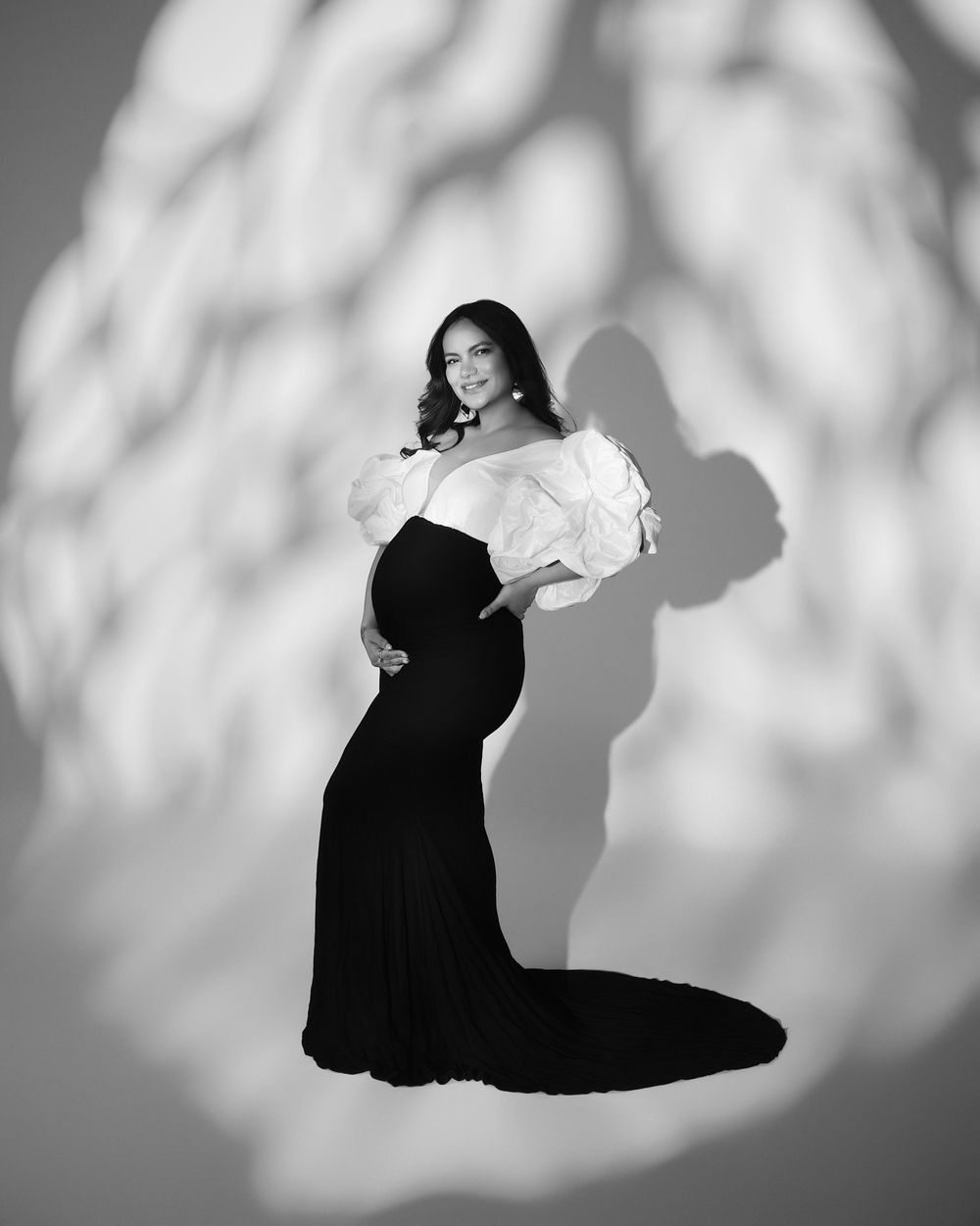 Black and white maternity photo of elegant pregnant woman in off-shoulder white top and black gown with dramatic shadow backdrop.