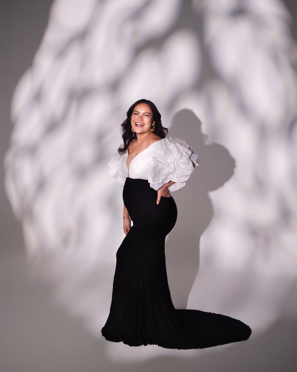 Pregnant woman in elegant black and white gown with ruffled sleeves, posing against a soft shadowed backdrop.