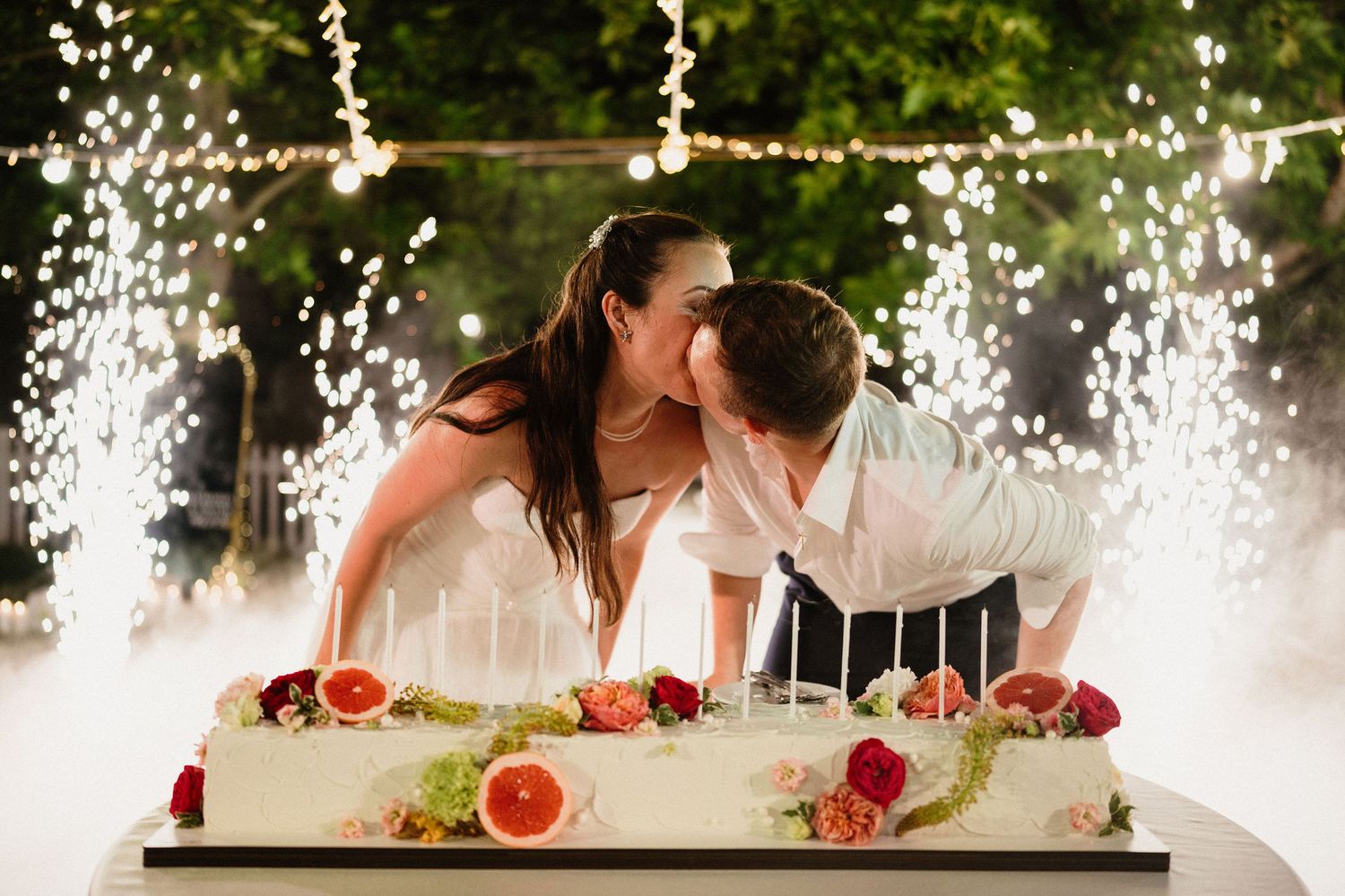Bride and groom sharing a kiss over a decorated wedding cake with candles and fruit, surrounded by sparkling lights.