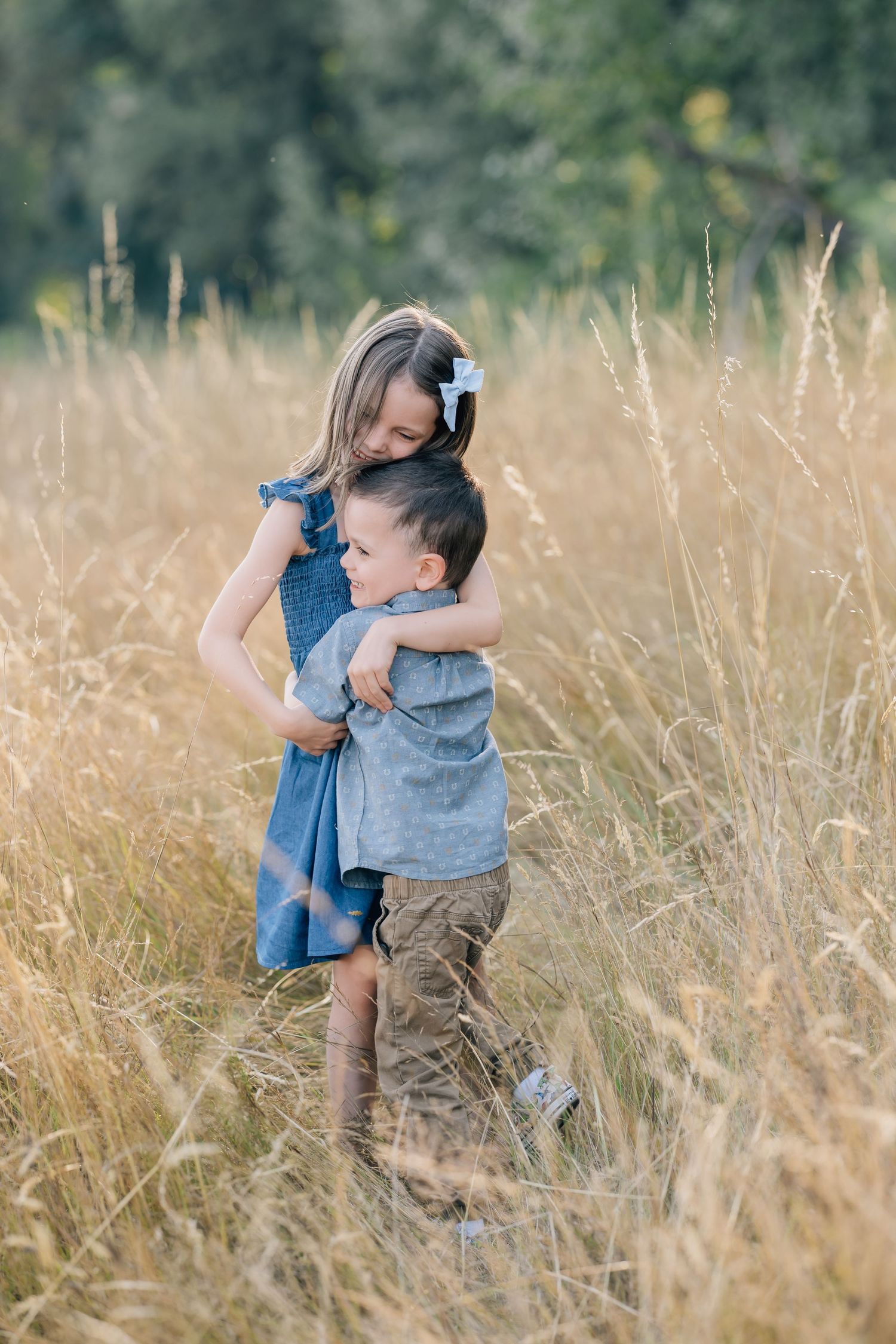 Two young siblings share a tender hug in a golden field, girl in blue dress with hair bow embracing little boy.