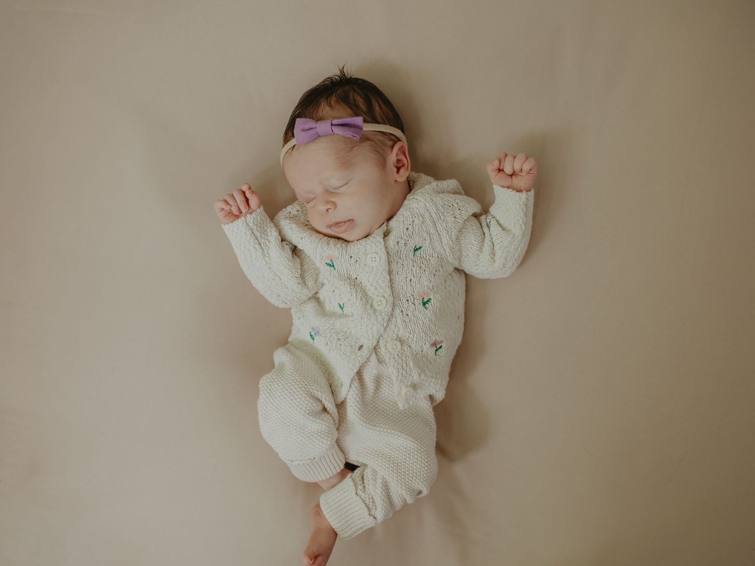 Sleeping newborn baby in cream knit romper with purple floral headband lying on beige background.