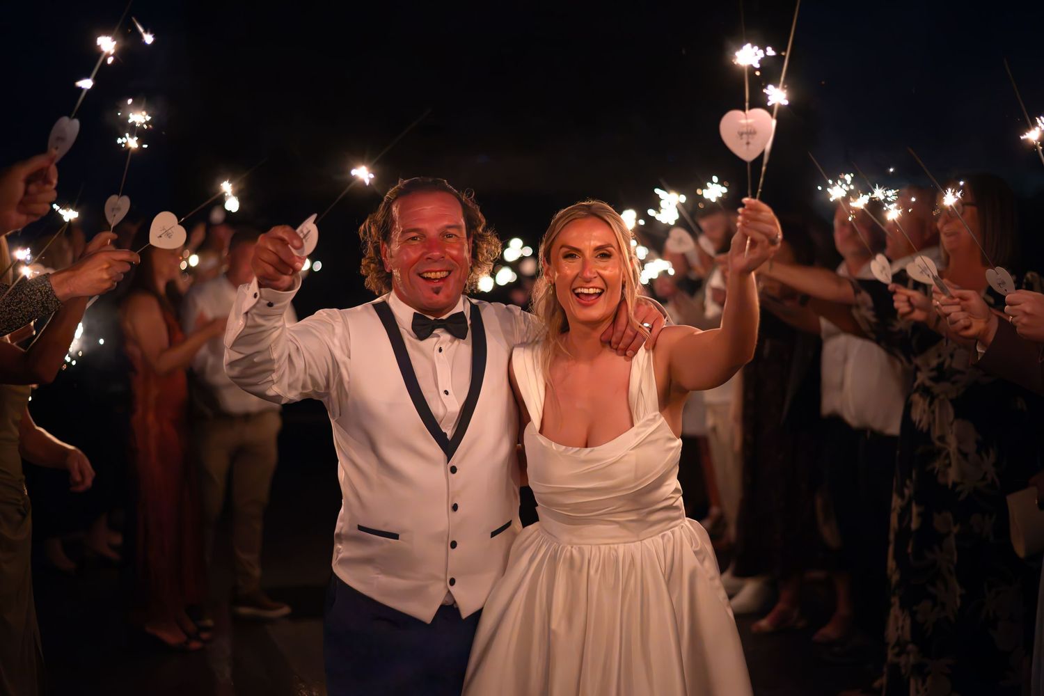 Newlywed couple smiling and holding sparklers surrounded by wedding guests during a nighttime sparkler send-off celebration.