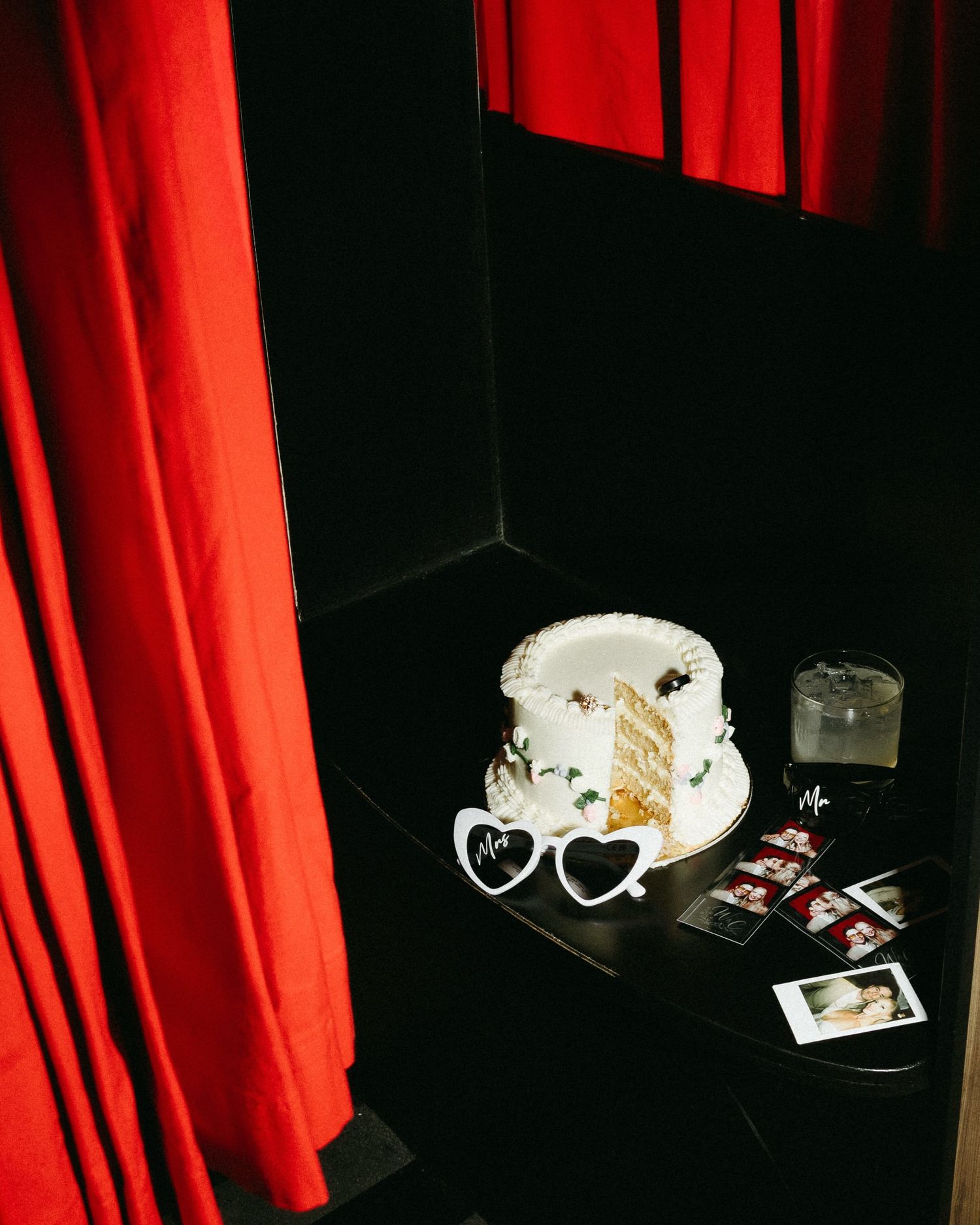 White wedding cake with heart-shaped sunglasses, Polaroid photos, and red curtain backdrop on dark table taken by Melissa Choi photography
