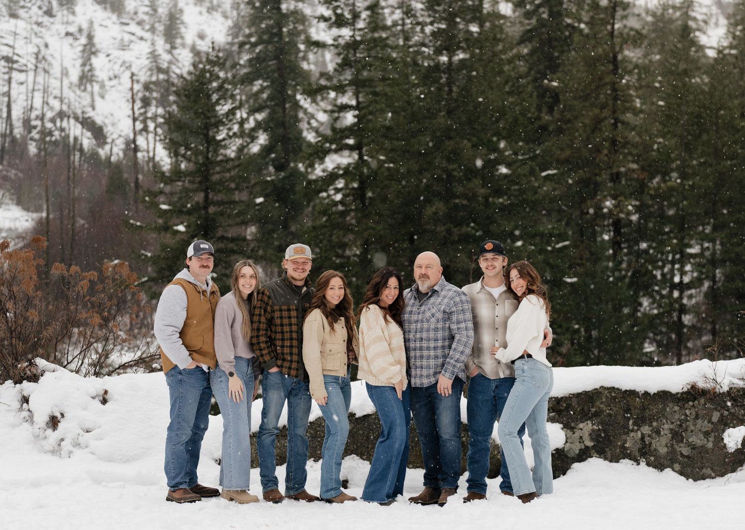 Family of seven posing together in snowy mountain landscape surrounded by pine trees during winter.