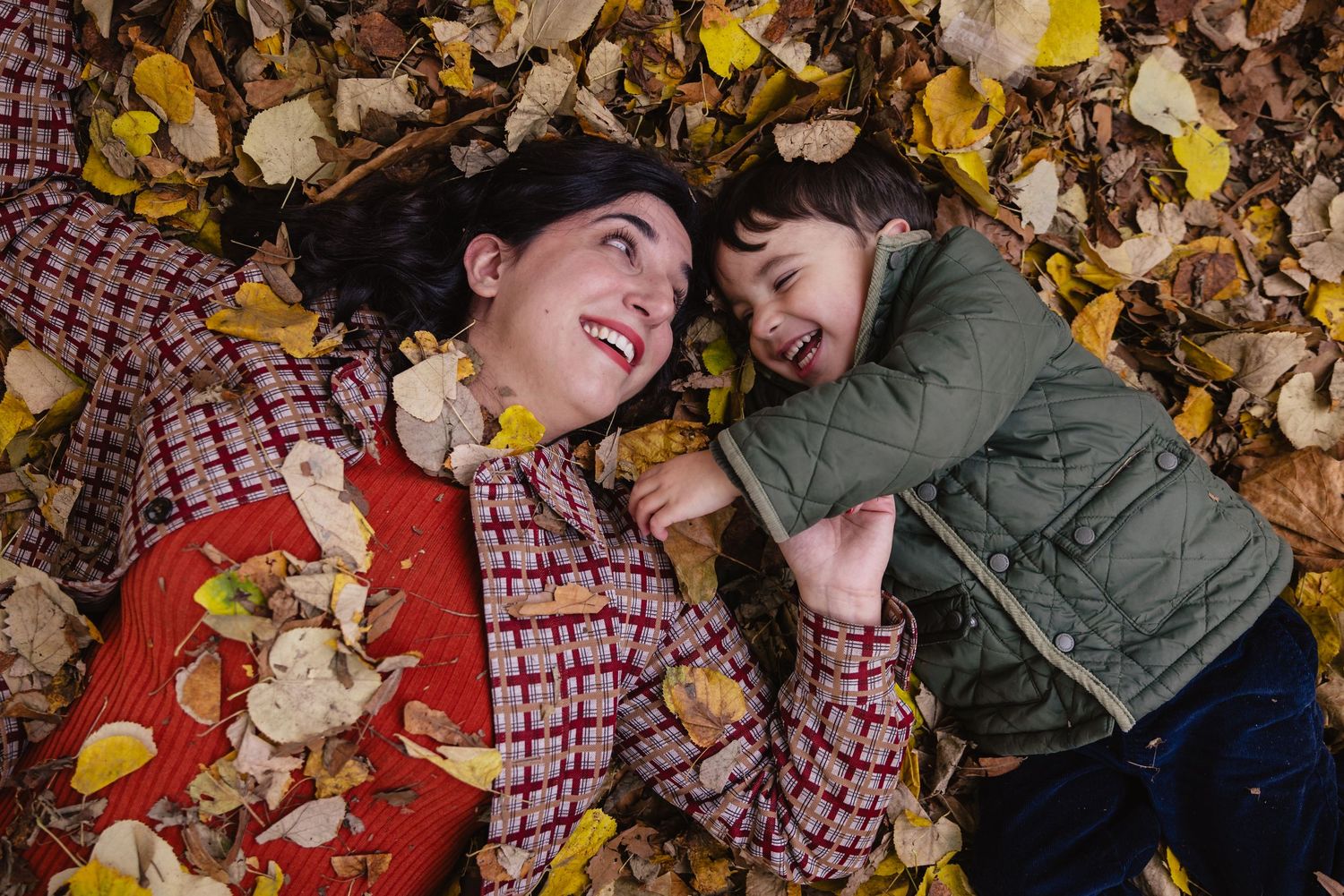 Une mère et son fils se regardent en riant, allongés sur un tas de feuilles mortes en automne