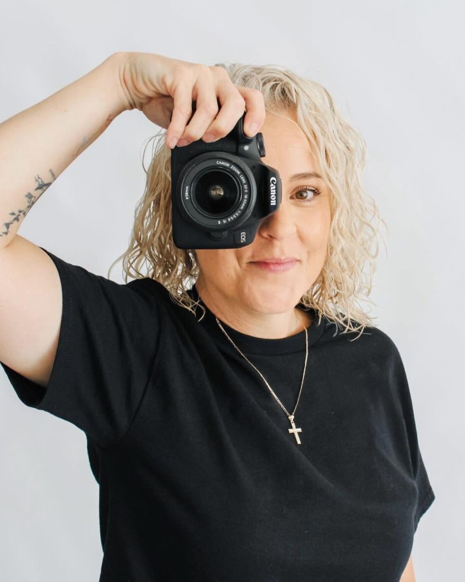 Smiling blonde woman in a black t-shirt and cross necklace holds a Canon camera up to her eye against a plain light backdrop.