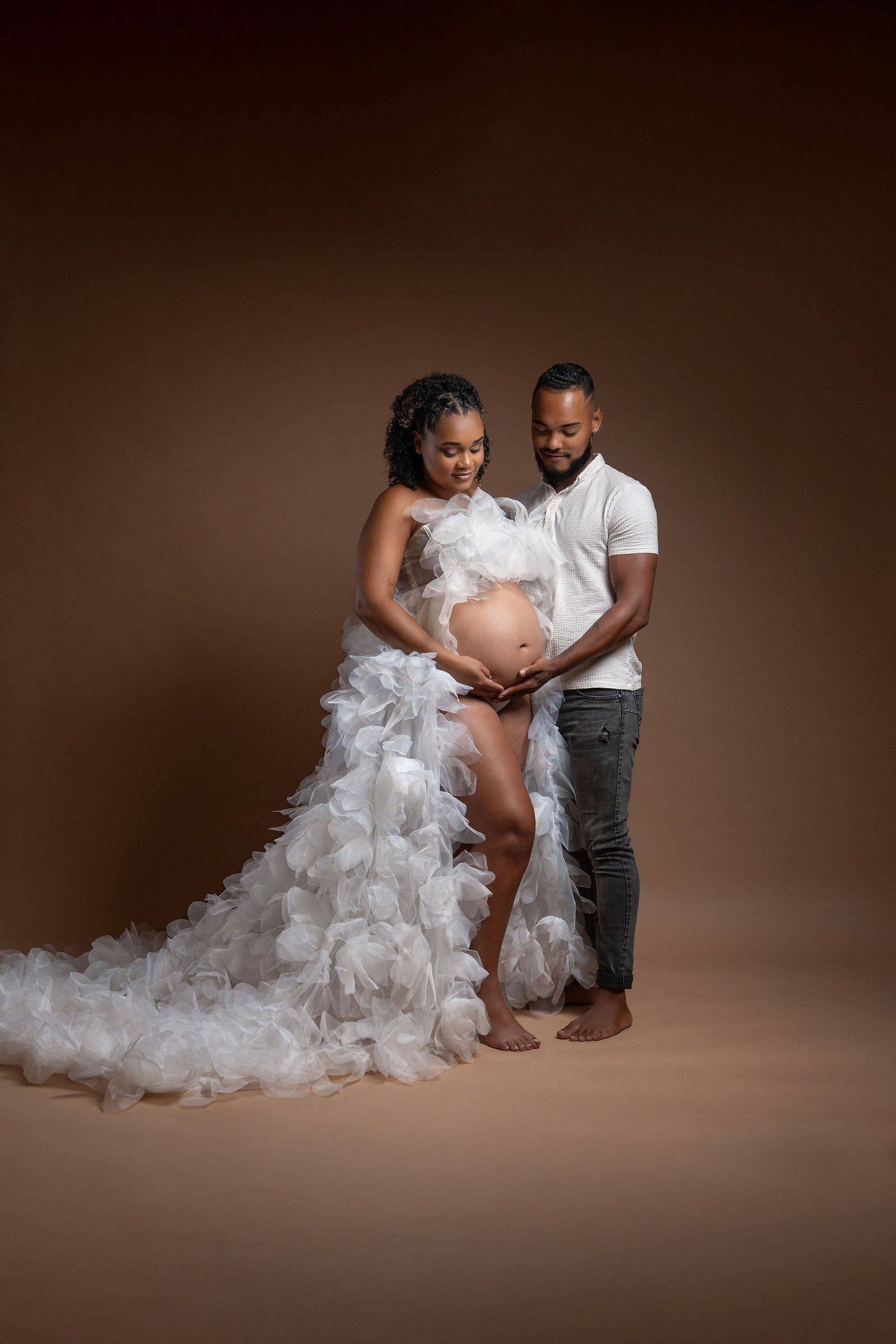 Pregnant woman in white ruffled gown with partner admiring baby bump during maternity photoshoot on brown background.