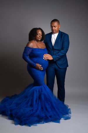 Pregnant woman in royal blue lace mermaid gown with partner in blue suit posing for maternity photo.