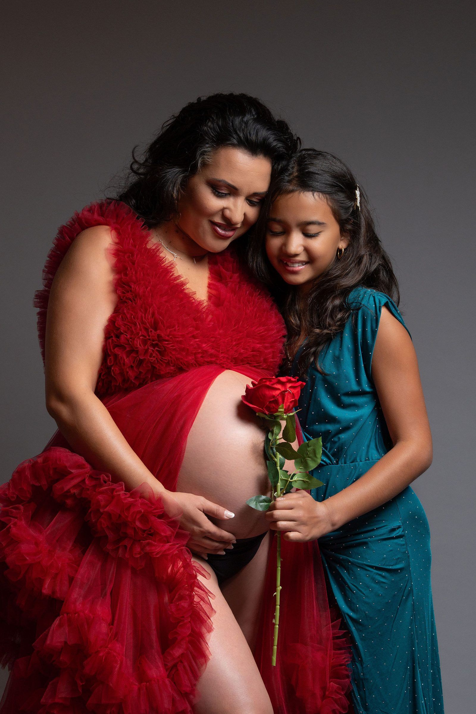 Pregnant woman in red tulle gown with daughter in teal dress holding a red rose, studio maternity photo.