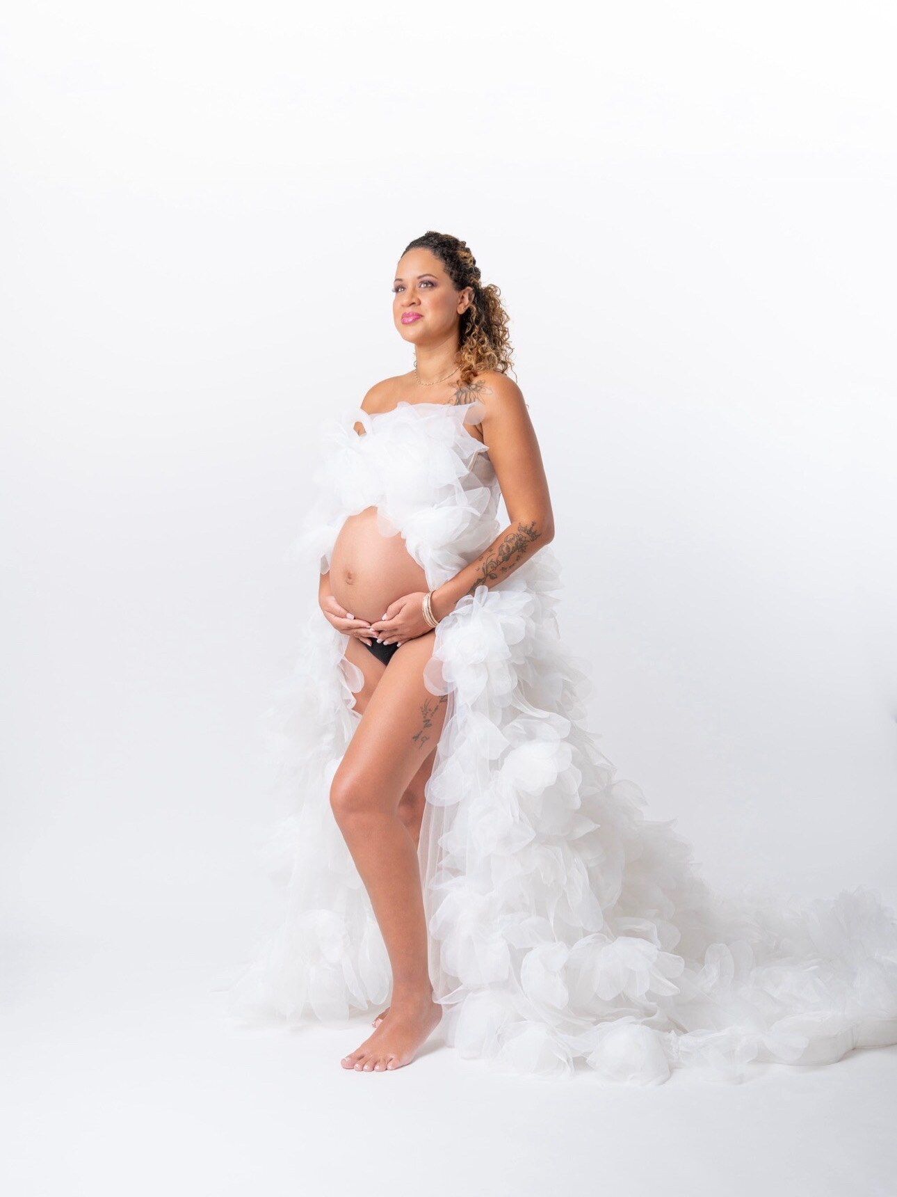 Pregnant woman in white ruffled maternity gown posing gracefully in a studio with white background.