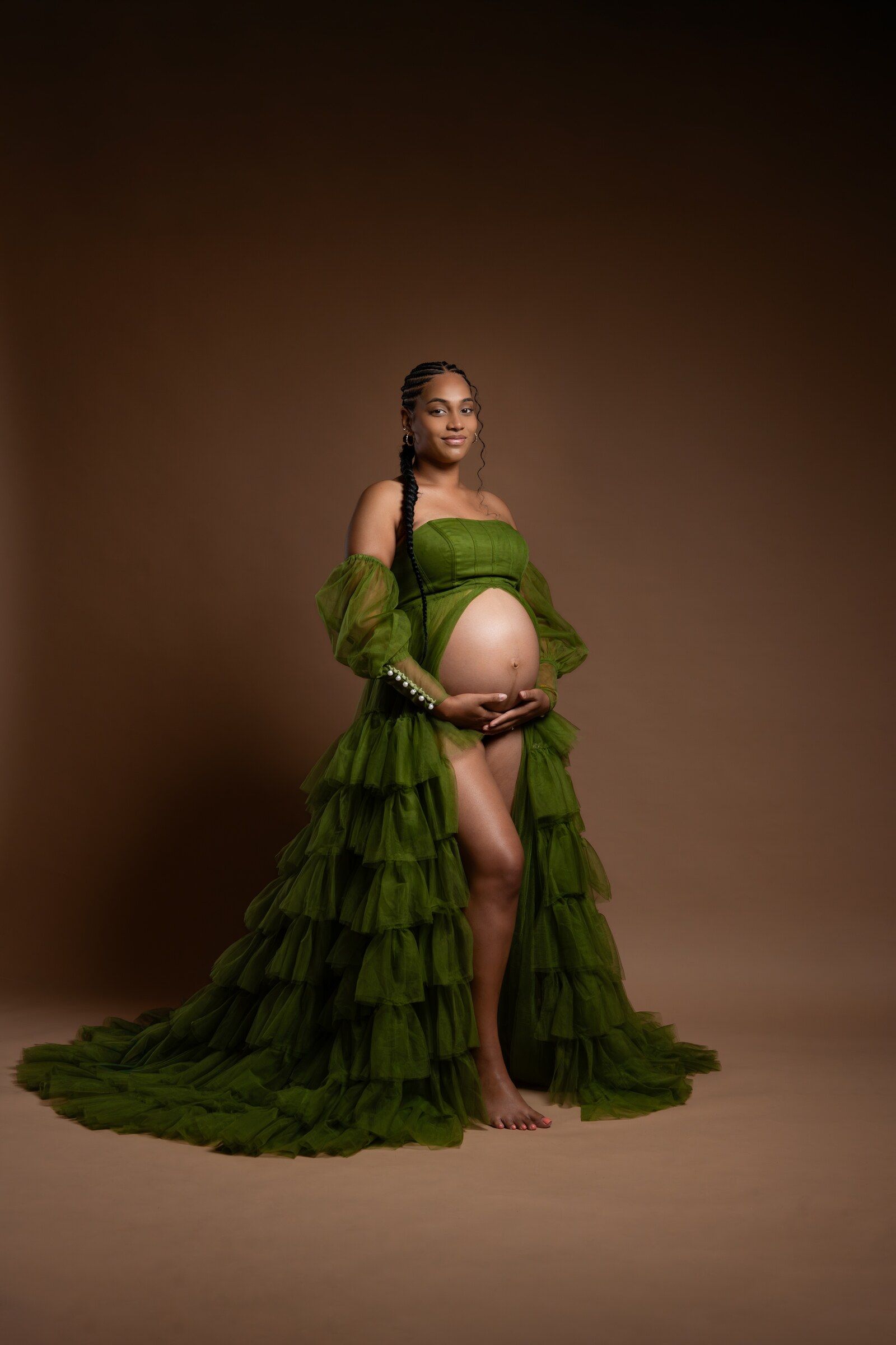 Pregnant woman in green ruffled tulle maternity gown cradling baby bump against brown studio background.