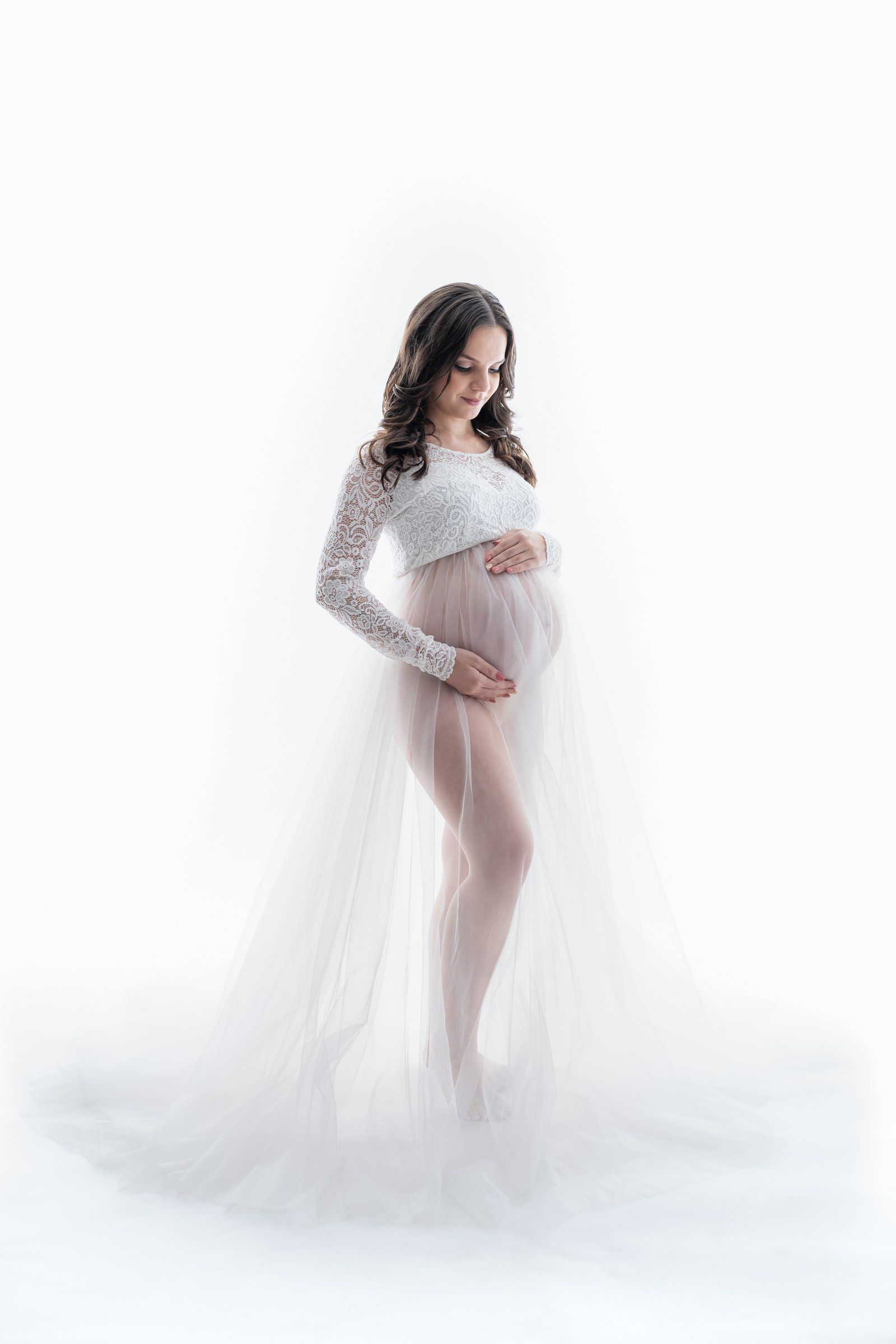Pregnant woman in sheer white lace maternity gown cradling baby bump against white background.
