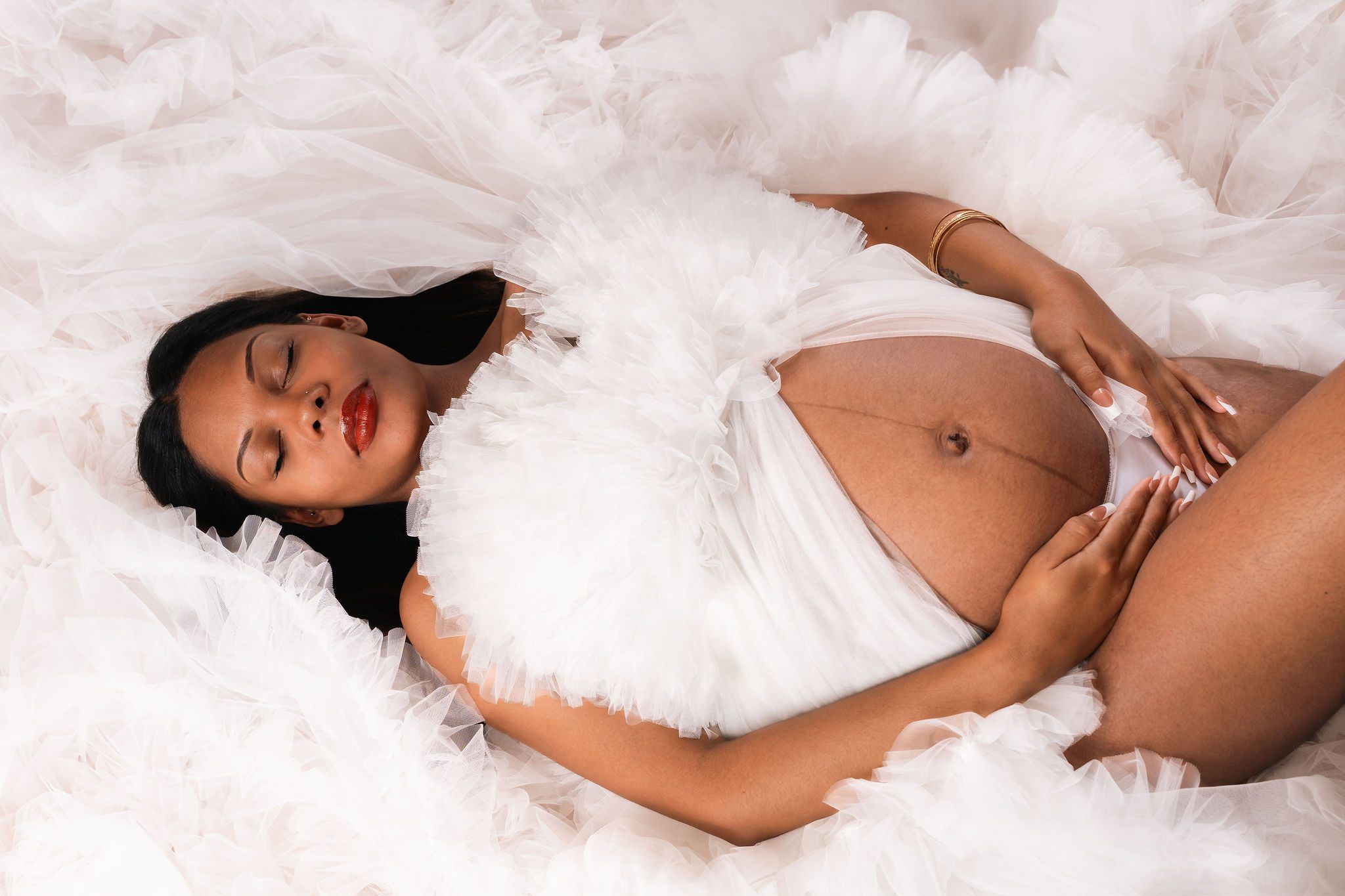 Pregnant woman lying on white fluffy fabric, cradling her bare baby bump in a serene maternity photo shoot.