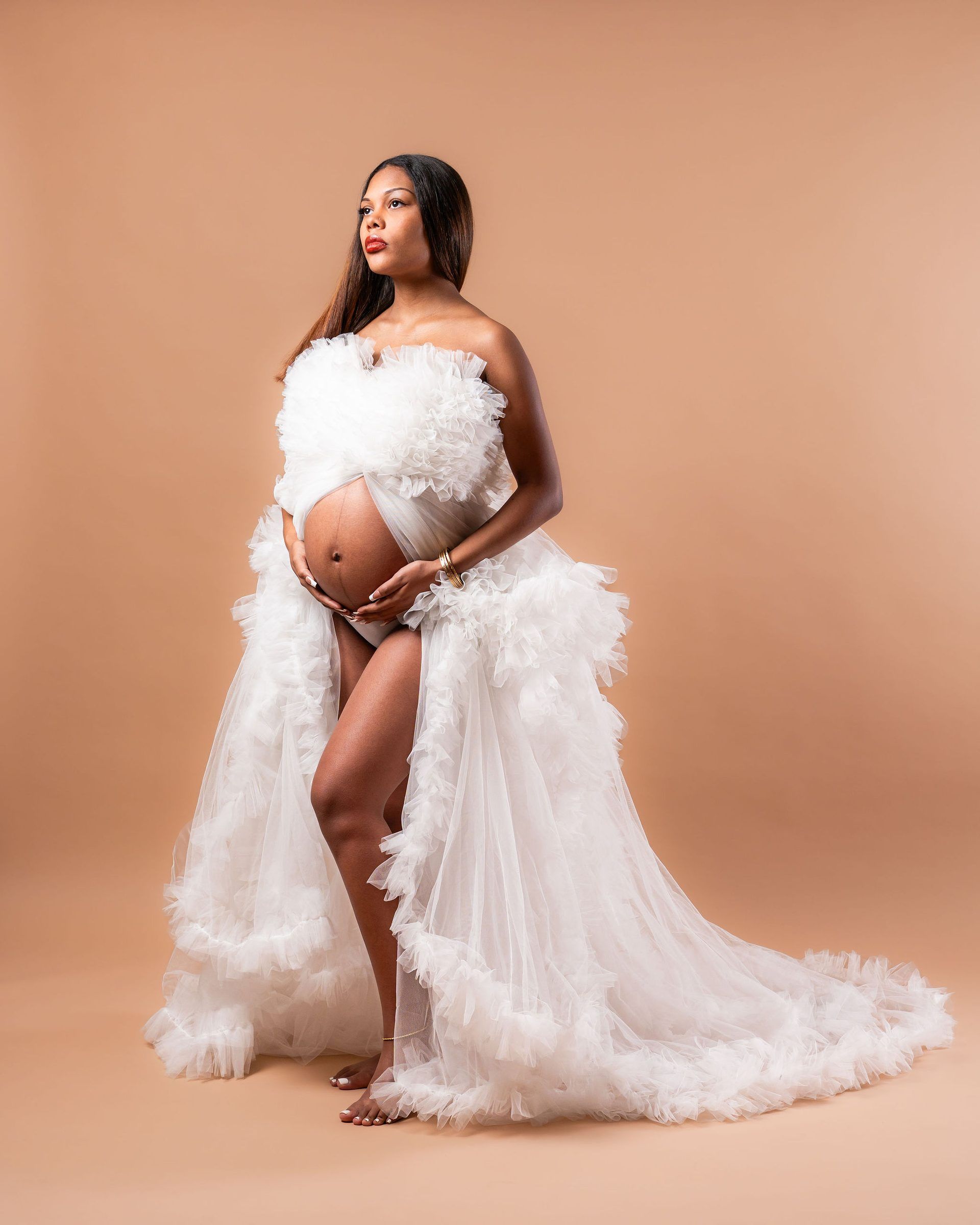 Pregnant woman in white feathered tulle maternity gown posing elegantly against a warm beige studio background.