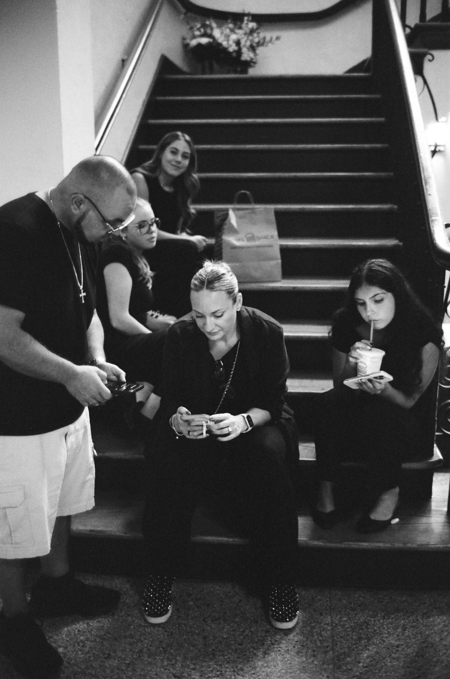 Black and white photo of a group of people sitting on stairs, looking at phones and drinking beverages indoors.