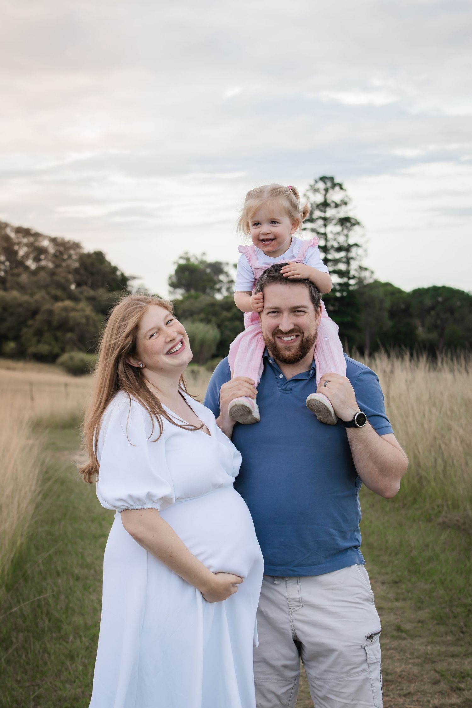 Pregnant woman in white dress smiles with husband and toddler on shoulders during maternity photoshoot in a scenic field in Rouse Hill.