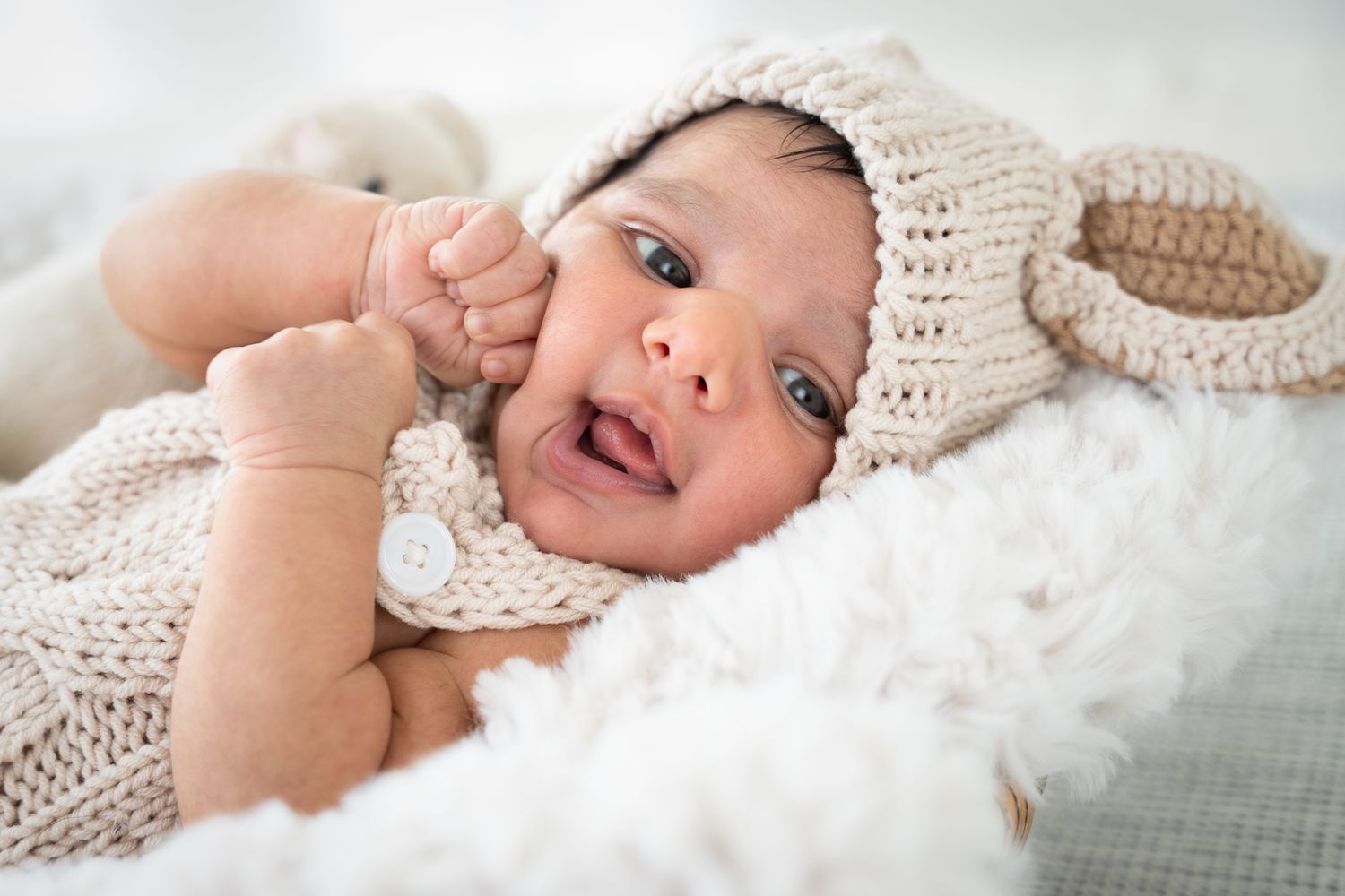 Awake newborn wearing a cream knitted outfit and floppy-eared hat, lying on a soft white blanket.