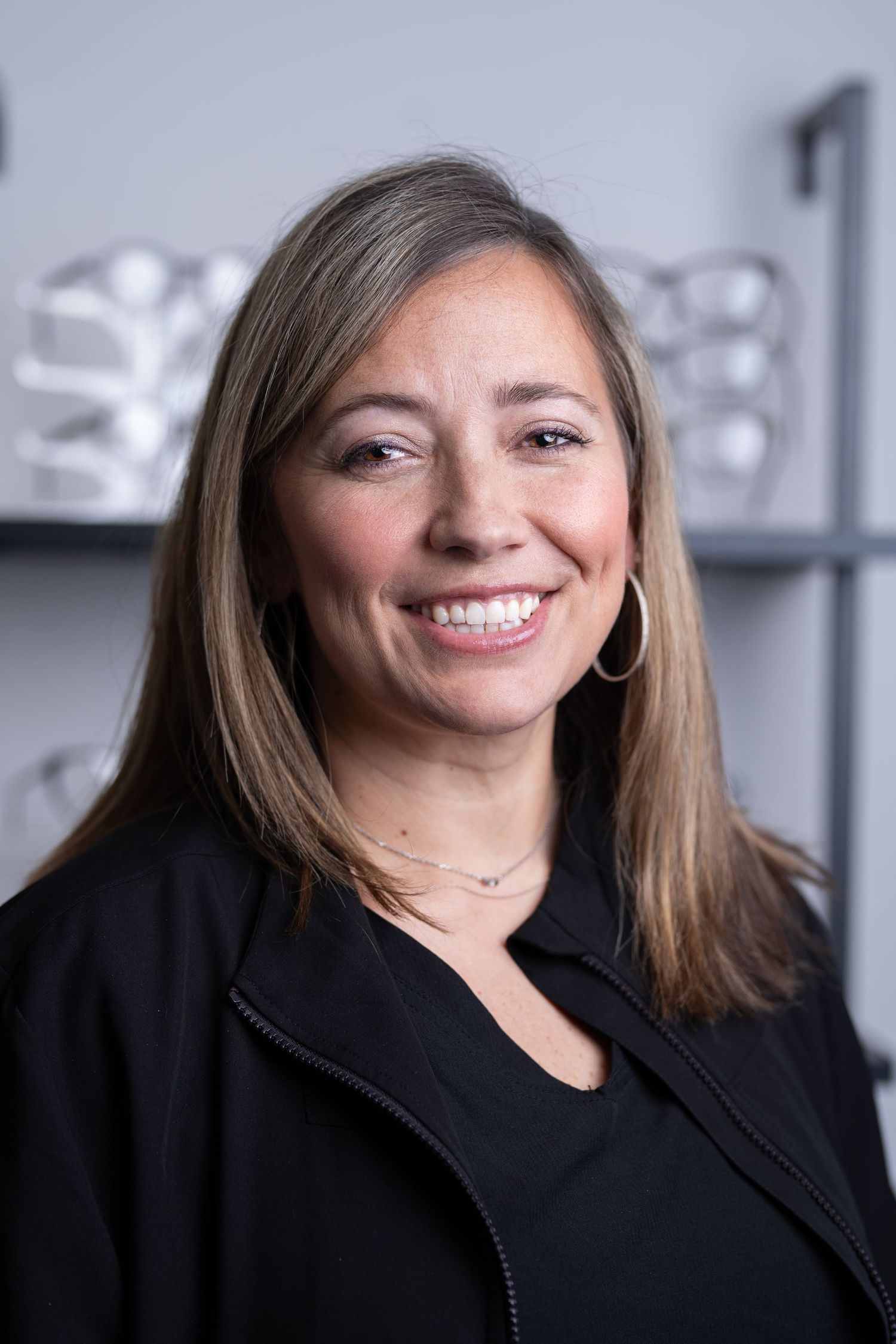 Smiling professional woman with brown hair wearing black jacket in front of gray shelving display.
