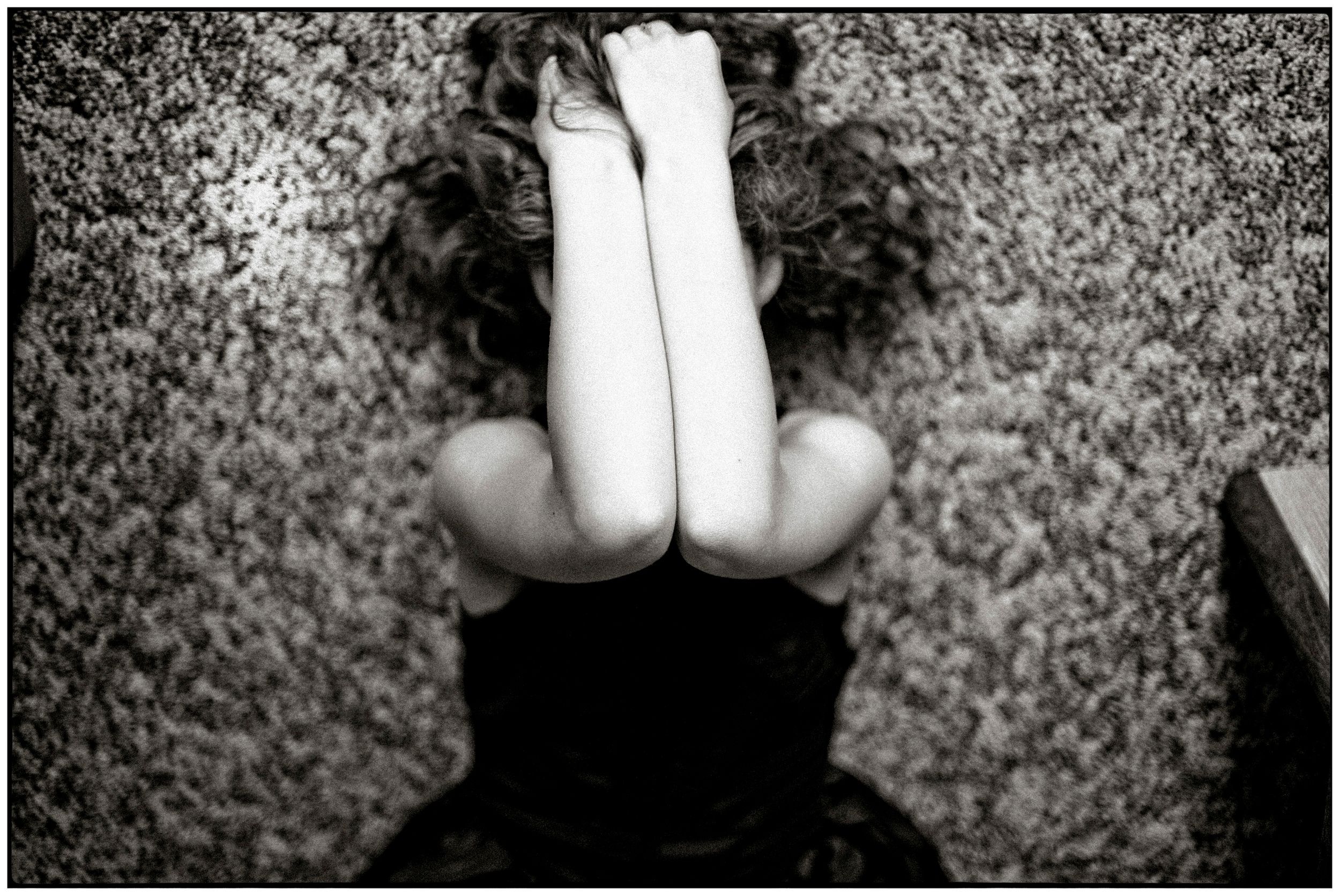 Black and white photo of a curly-haired person sitting on ground, hugging knees to chest in a contemplative pose.