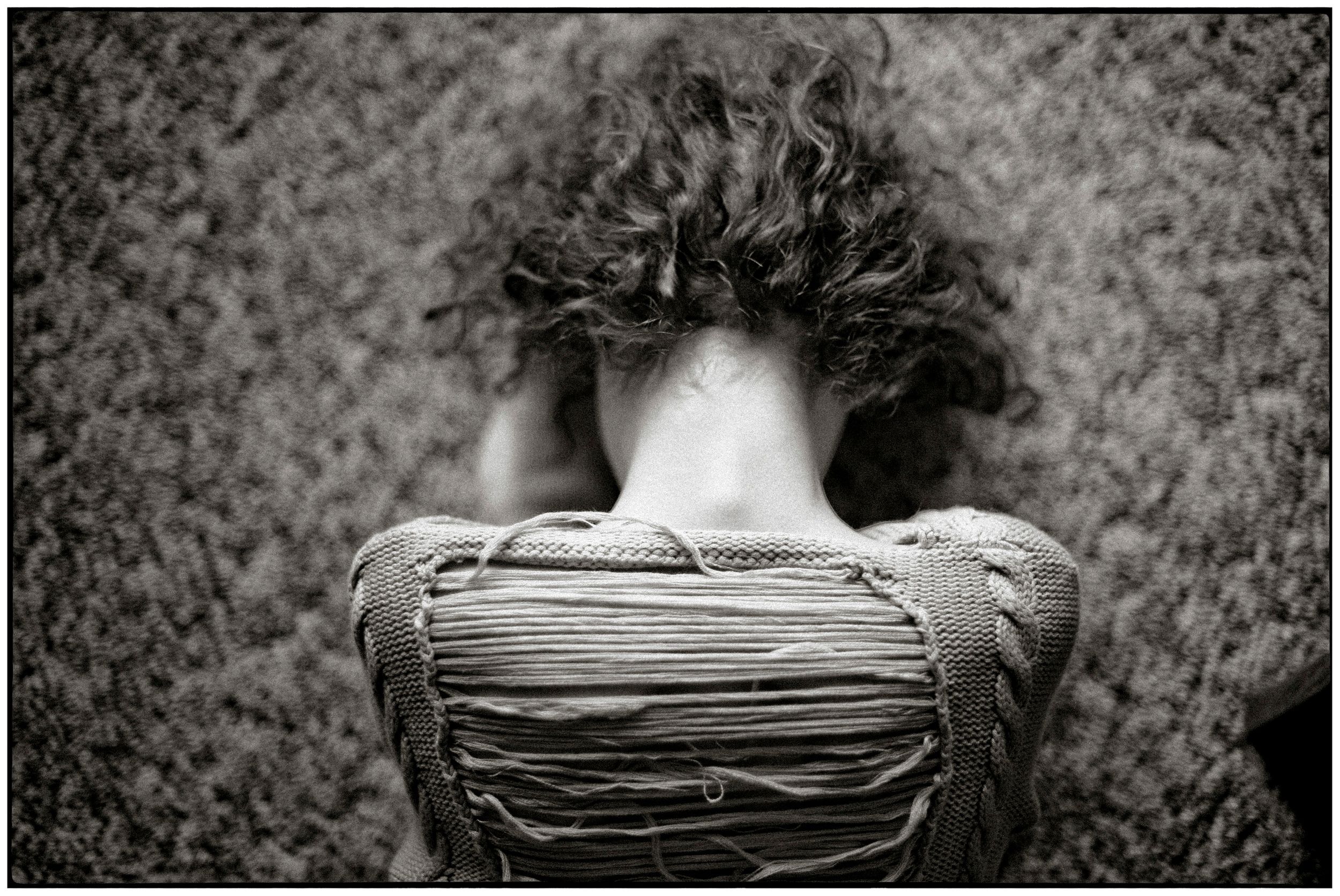 Black and white photo of a person with curly hair sitting hunched over a worn backpack on grass.