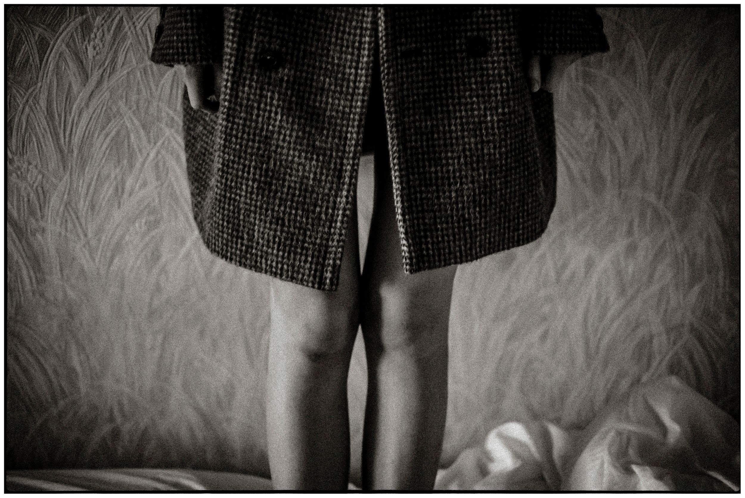 Black and white photo of a person standing in a coat, showing bare legs against a textured wallpaper background.