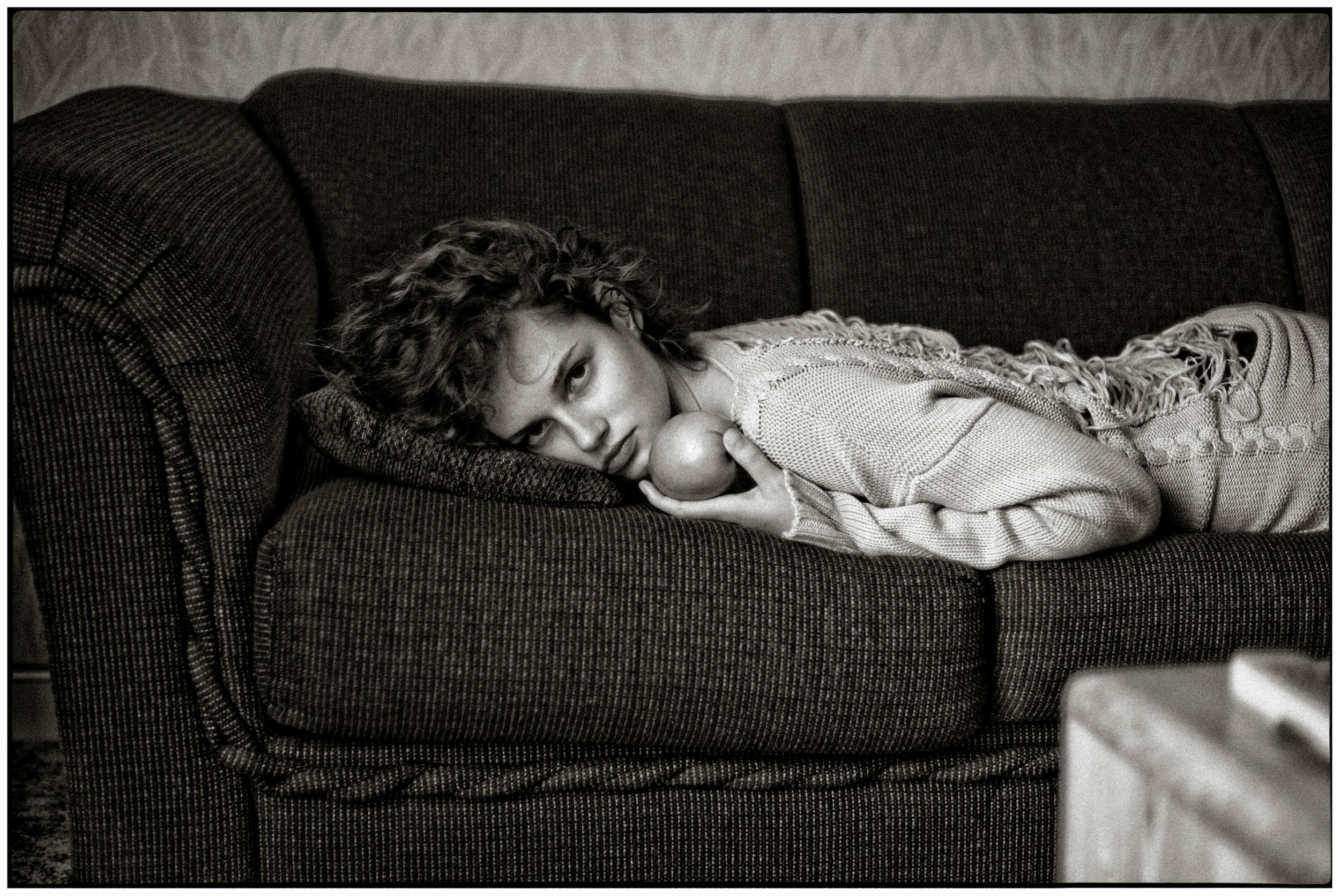 Black and white photo of a young person lying on a dark sofa, looking contemplative in casual clothing.