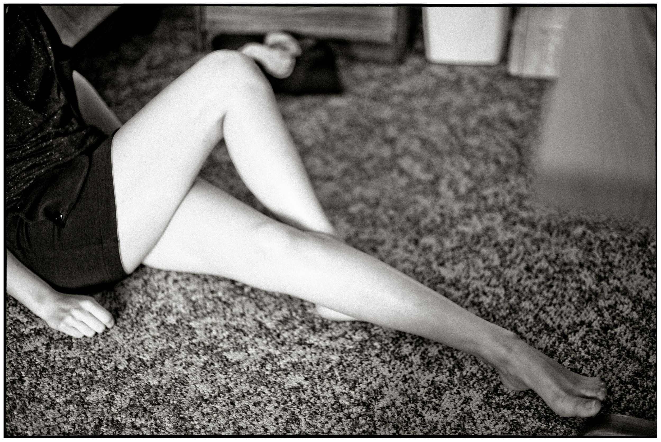 Black and white photo of a woman sitting on a carpet, showcasing her bare legs in a short dark skirt.
