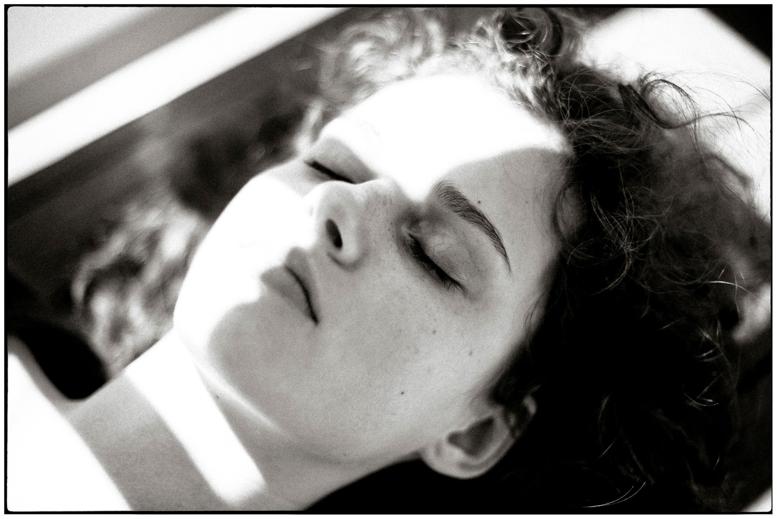 Black and white portrait of a young woman with curly hair, eyes closed, bathed in soft sunlight.