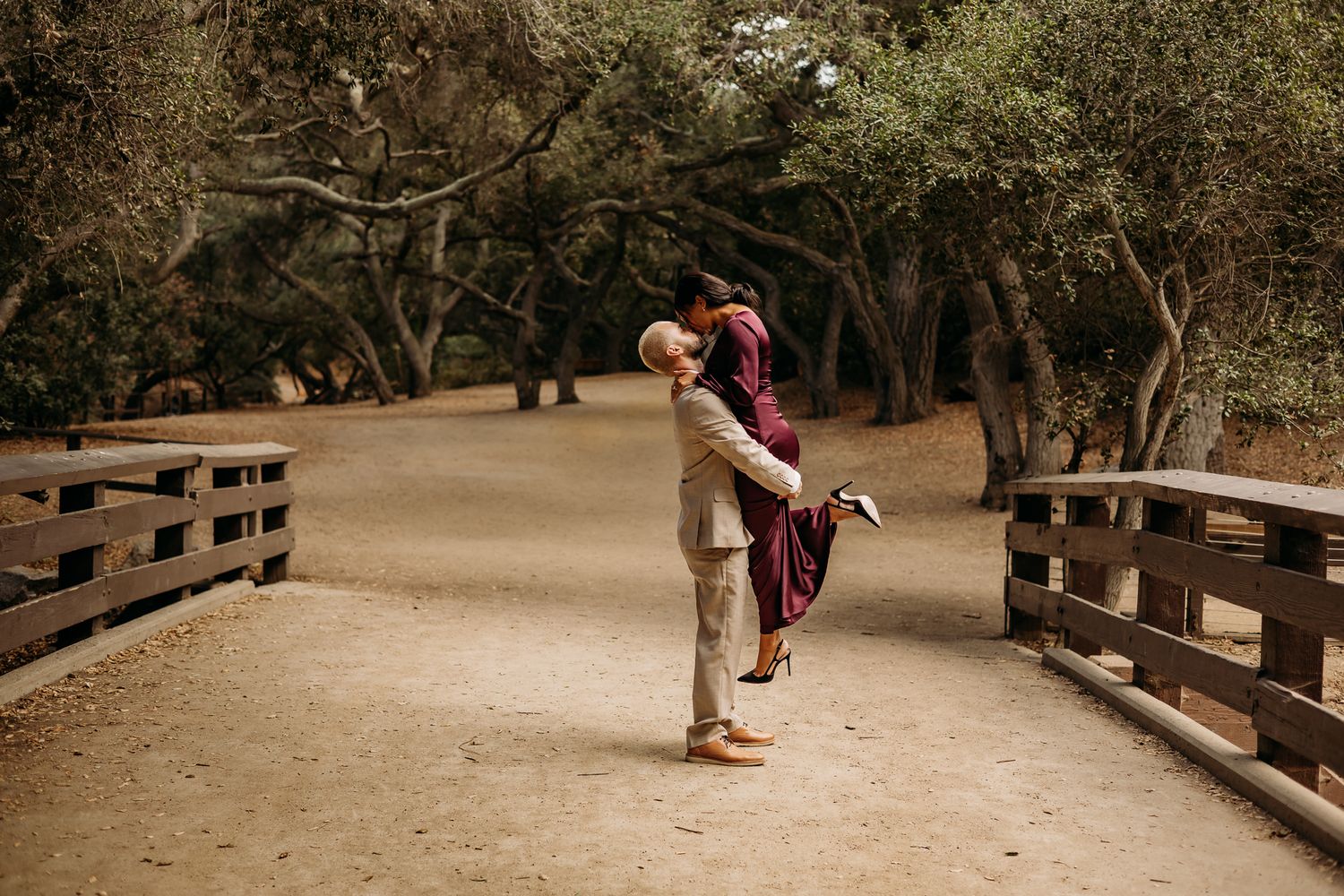 A groom lifts his bride in a maroon dress on a wooden bridge path surrounded by lush green trees.
