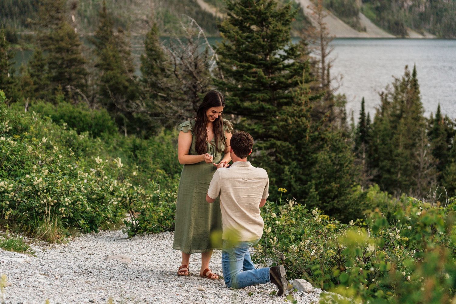 Man proposing to woman on a scenic mountain trail with evergreen trees and a lake in the background.