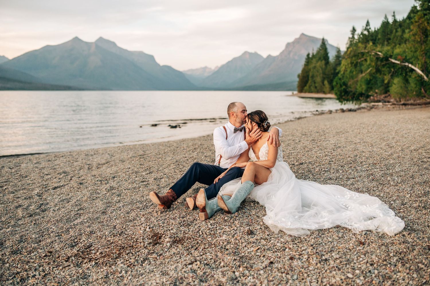 Couple in wedding attire sitting on rocky beach with mountain lake and pine trees backdrop at sunset.