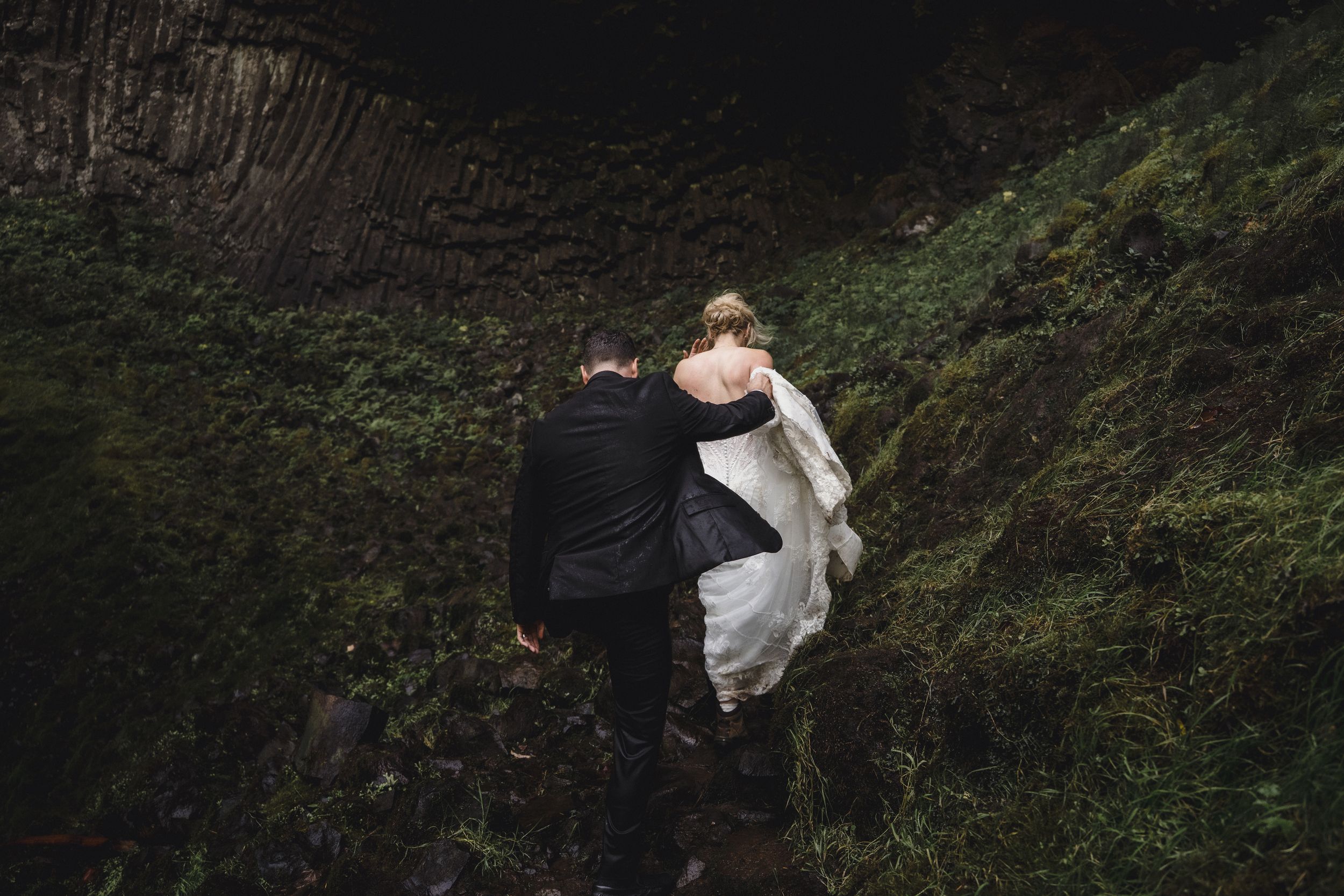 A bride and groom walk hand in hand up a lush green hillside during their outdoor wedding elopement.