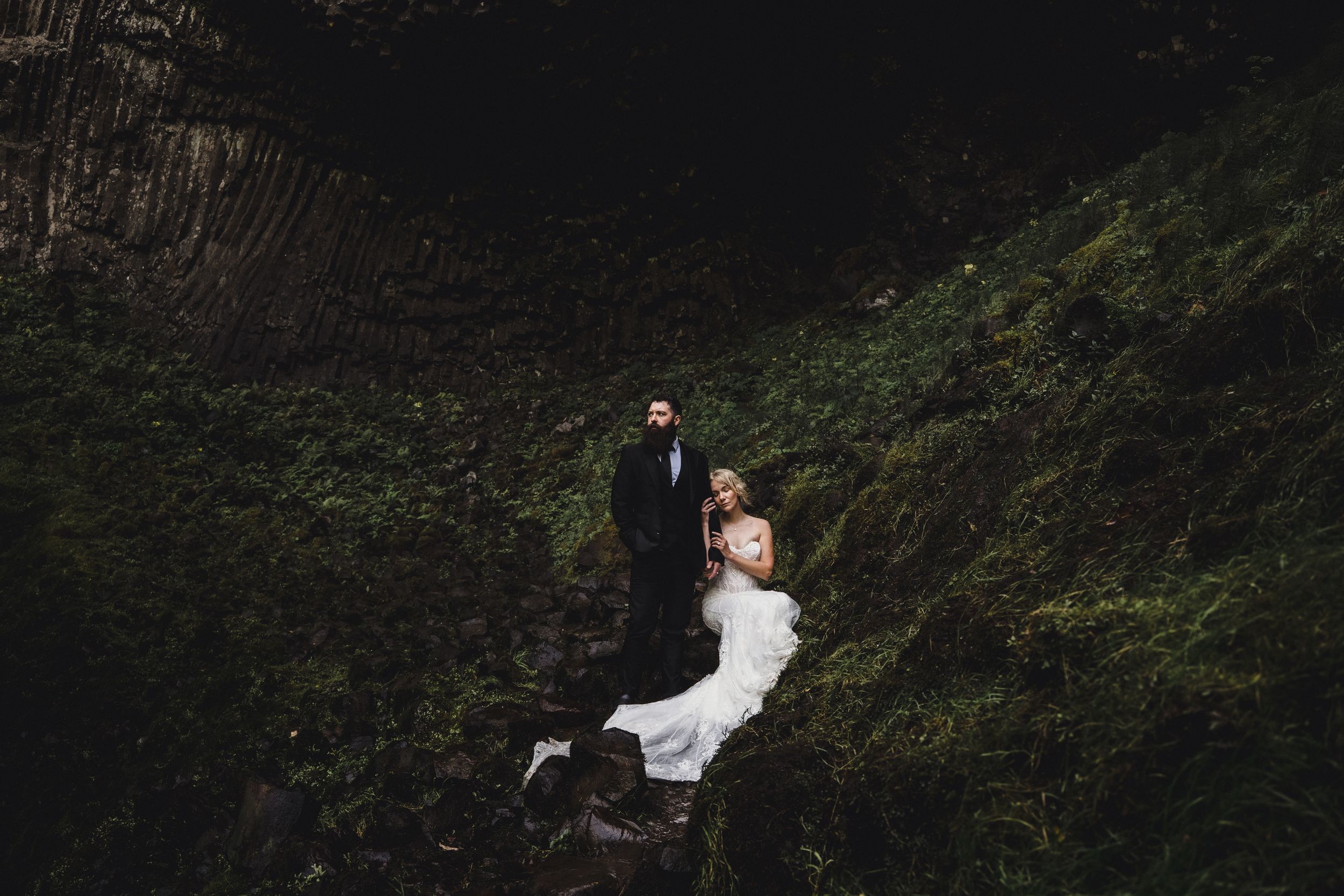 Bride and groom pose romantically on rocky cliff surrounded by lush green moss and foliage during outdoor wedding shoot.