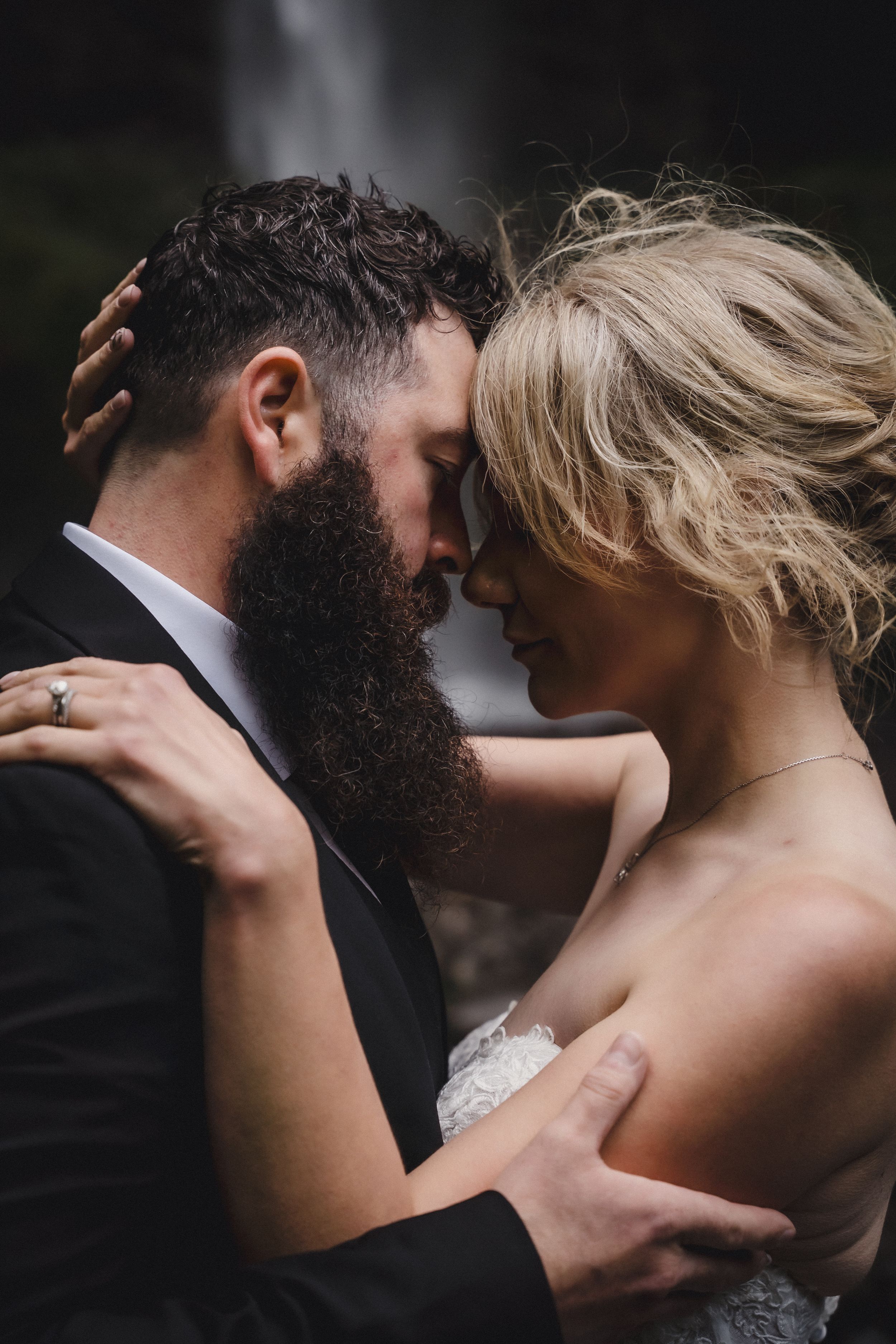 A bearded groom in a black suit and blonde bride in lace embrace tenderly with foreheads touching near a waterfall.
