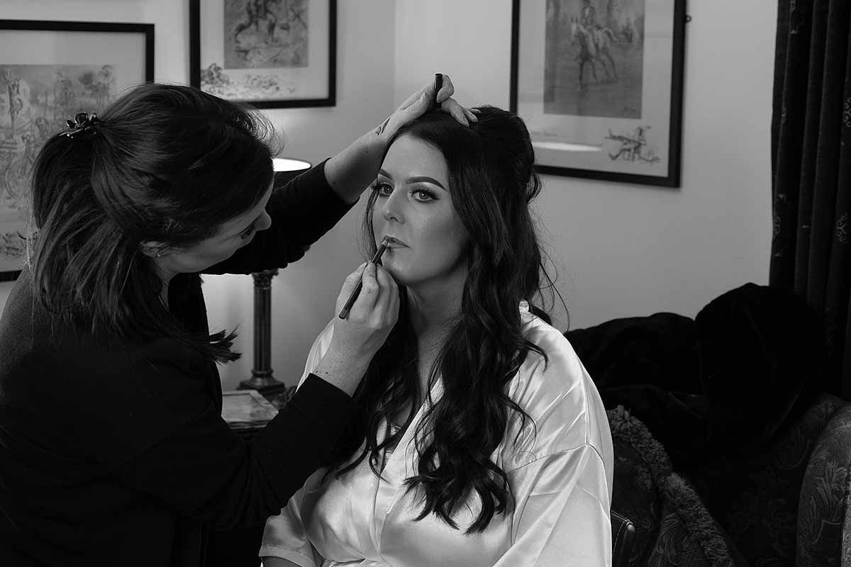 Black and white photo of a makeup artist applying lipstick to a bride during bridal prep at New House Farm wedding venue