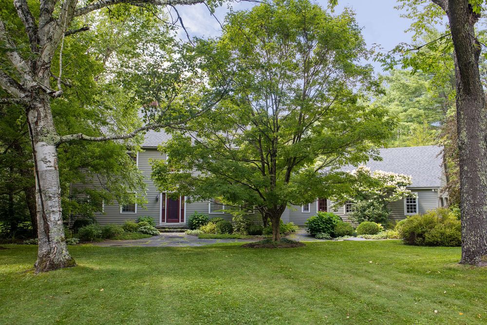 Charming gray colonial home surrounded by lush mature trees and well-manicured lawn in a serene setting.