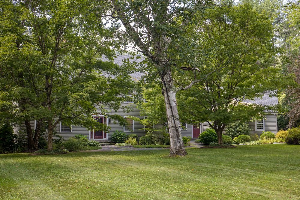 Classic gray colonial home nestled among towering mature trees with beautifully maintained green lawn.
