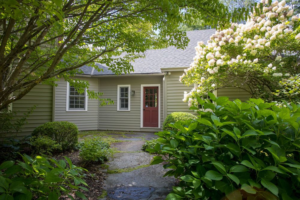 Sage green cape-style home with red door framed by blooming hydrangeas and lush garden landscaping.