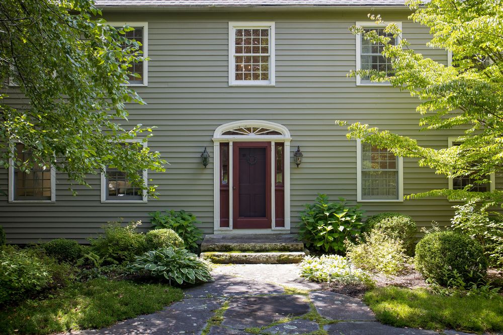 Elegant two-story gray colonial home featuring a decorative fanlight entry door surrounded by mature trees.